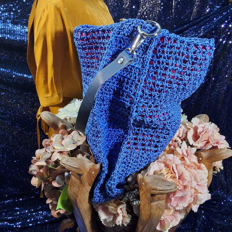 May include: A vibrant blue crocheted handbag with a red interior and a gray leather strap. The bag features a silver clasp and is displayed with a bouquet of pink and white flowers and a mustard-colored garment.