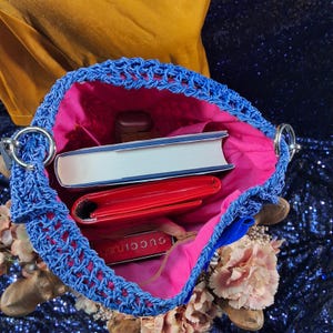 May include: A blue crocheted handbag with a pink interior. Inside the bag are a white book, a red wallet, and a small rectangular item with the text "GUCCIRUS". The bag has silver metal handles and is surrounded by flowers.
