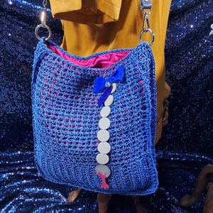 May include: A blue crocheted tote bag with a pink interior. The bag features a blue bow and a decorative chain of white circular beads. The bag has silver metal hardware and a gray strap.