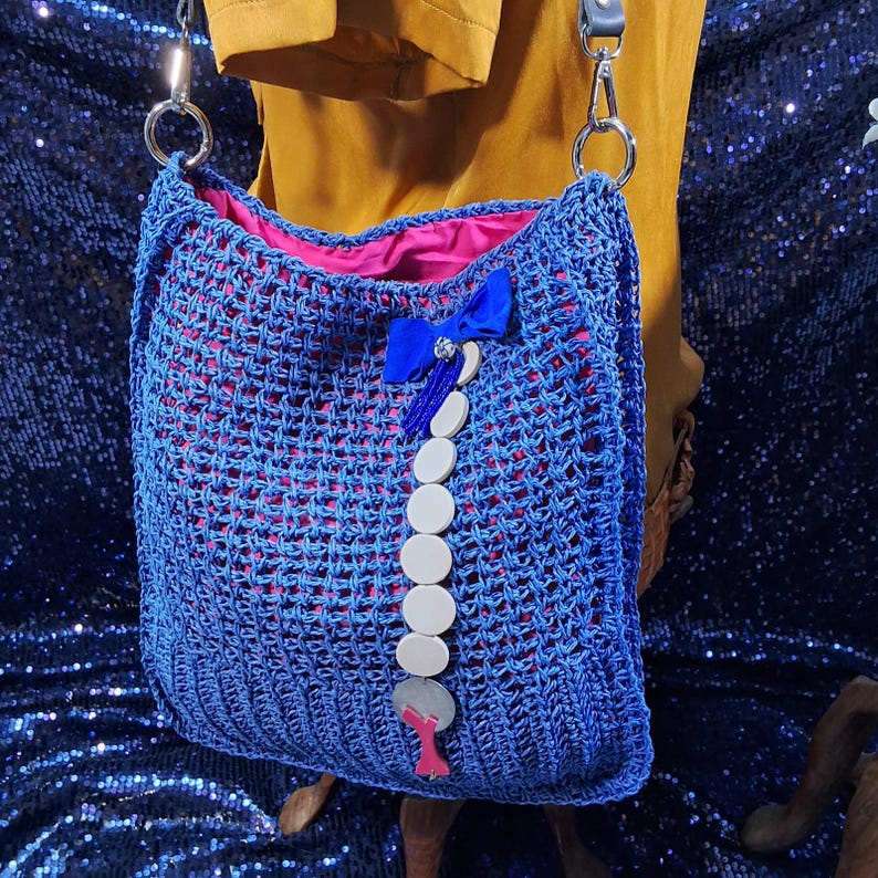May include: A blue crocheted tote bag with a pink interior. The bag features a blue bow and a decorative chain of white circular beads. The bag has silver metal hardware and a gray strap.