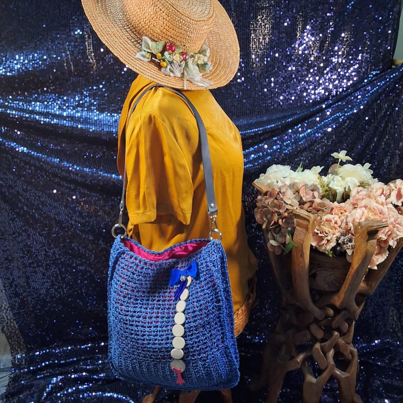 May include: A blue crocheted tote bag with a pink interior and a decorative blue bow. The bag has a gray strap and a series of white circular embellishments. A straw hat with floral accents is also visible.