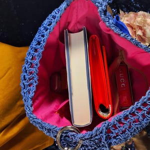 May include: A blue woven handbag with a pink interior. Inside the bag are a white book, a red notebook, and a small tag with the word "Gucci" on it. The bag has a silver metal ring.