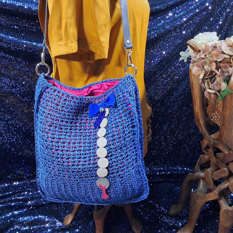 May include: A blue crocheted shoulder bag with a pink interior. The bag features a decorative blue bow and a string of white circular embellishments. The bag has a gray leather strap and silver metal rings.