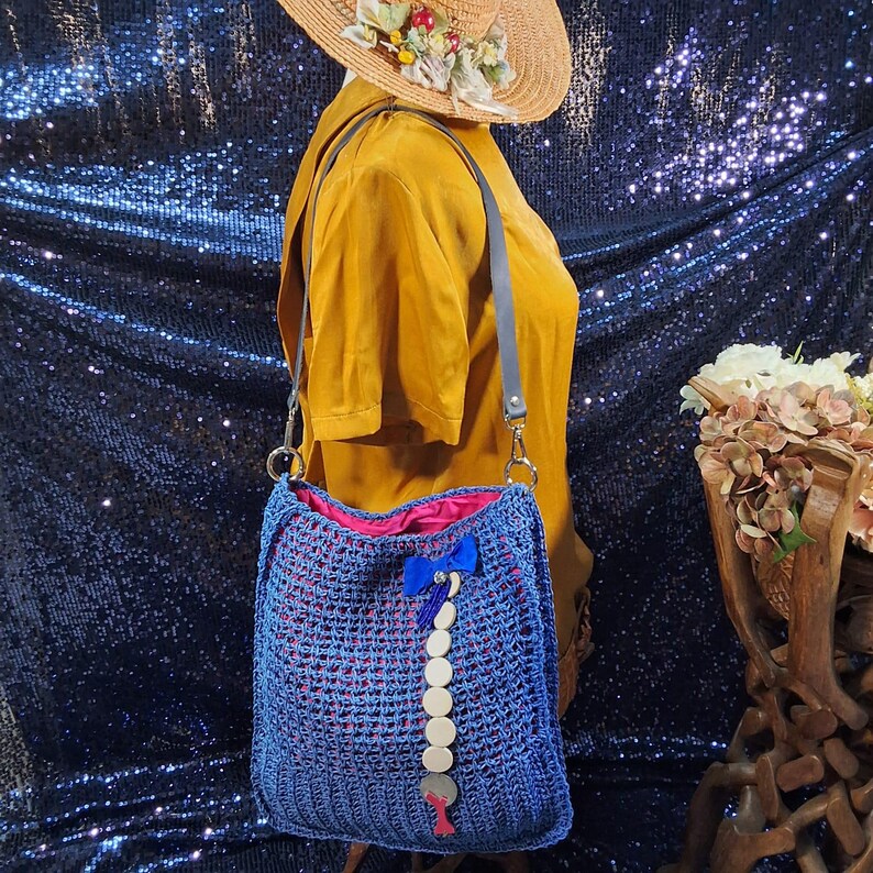 May include: A blue crocheted shoulder bag with a pink interior lining. The bag features a decorative blue bow and a string of wooden beads. The bag has a gray strap and is displayed with a straw hat.