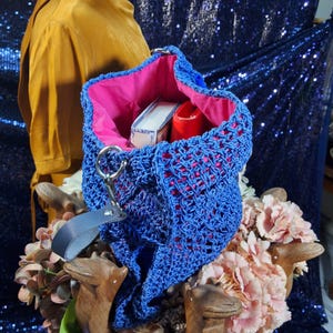 May include: A blue crocheted tote bag with a pink interior. The open weave bag has a silver ring handle and a black leather strap. Inside the bag are a white box and a red item. The bag is displayed with pink flowers.