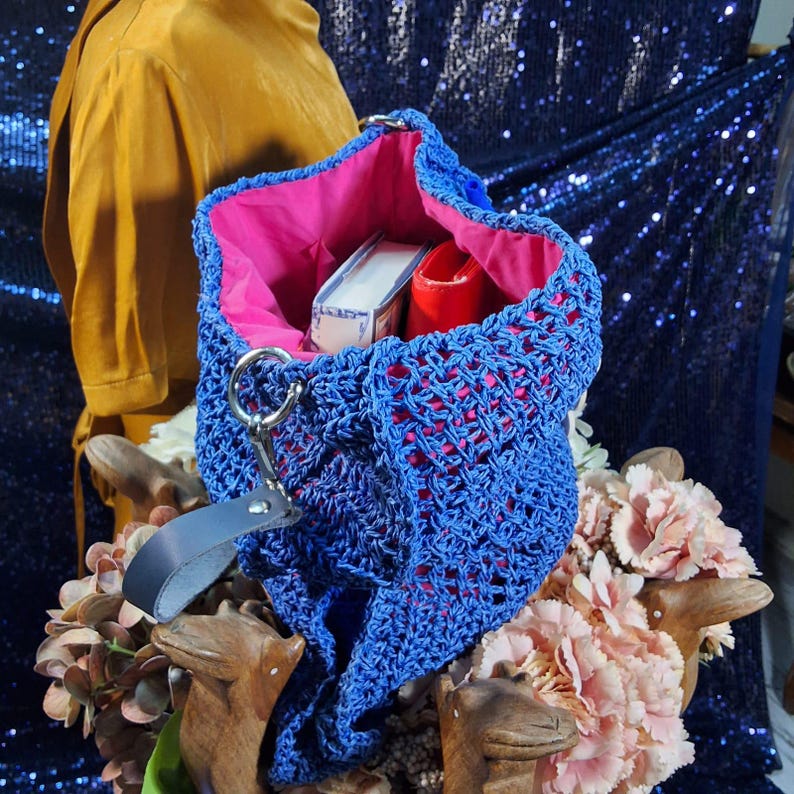 May include: A blue crocheted tote bag with a pink interior. The open weave bag has a silver ring handle and a black leather strap. Inside the bag are a white box and a red item. The bag is displayed with pink flowers.