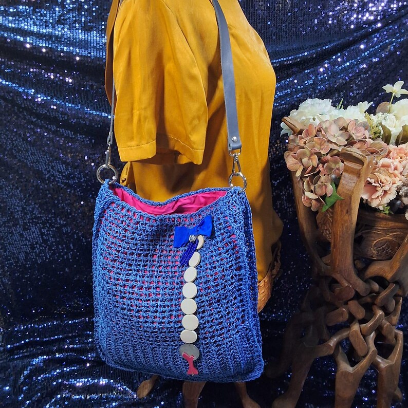 May include: A blue crocheted shoulder bag with a pink interior lining. The bag features a blue bow and a decorative strand of white and silver embellishments. It has a gray leather strap and silver hardware.