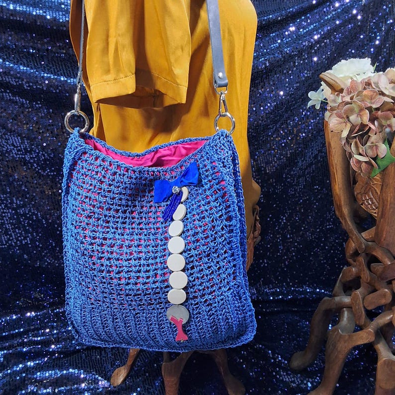 May include: A blue crocheted shoulder bag with a pink interior lining. The bag features a blue bow and a decorative strand of white and gray circular embellishments. The bag has a gray leather strap and silver metal hardware.