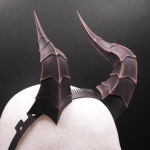 Puede incluir: Un par de cuernos de disfraz marrones oscuros y negros sujetos a una diadema negra. Los cuernos son curvos y texturizados, con un diseño en capas. La diadema se lleva en una cabeza de maniquí blanca sobre un fondo oscuro.