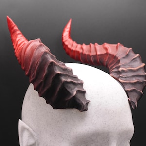 May include: A pair of devil horns, with a gradient of red to black, are mounted on a white head form. The horns have a textured, ridged appearance, and curve slightly. The horns are a costume accessory.