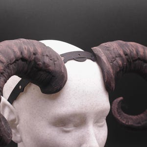 Puede incluir: Un par de cuernos grandes y curvados, de color marrón oscuro con una superficie texturizada, unidos a una diadema negra. Los cuernos están montados sobre una cabeza de maniquí blanca sobre un fondo negro. Accesorio de disfraz.