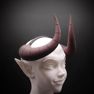 Puede incluir: Una cabeza blanca con orejas puntiagudas adornada con un par de cuernos curvos de color marrón oscuro. Los cuernos están segmentados y sujetos a la cabeza con una correa negra. Los cuernos son de color marrón oscuro sobre un fondo oscuro.