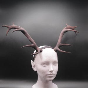 May include: A brown antler headband with a dark leather band. The antlers are detailed with multiple points and are attached to a white head form. The background is a dark gray.