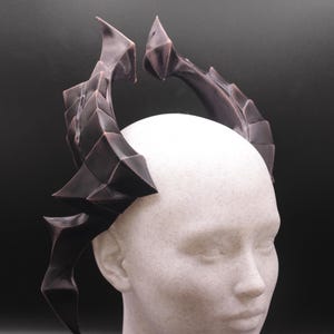 May include: A pair of dark brown, stylised horns, possibly for a costume or cosplay, are mounted on a white mannequin head. The horns have a textured, geometric design against a dark background.