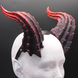 May include: A pair of devil horns, red at the tips and fading to black, attached to a black headband. The horns are textured and curved, with a spiked appearance. The horns are on a white mannequin head against a black background.