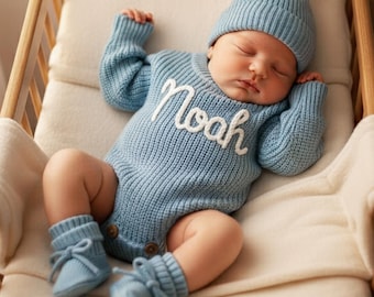 Może przedstawiać: Śpiące dziecko w jasnoniebieskim dzianinowym stroju, w tym czapka, sweter, skarpetki i body z białym haftem z imieniem "Noah". Dziecko leży w drewnianym łóżeczku z kremowym kocem.