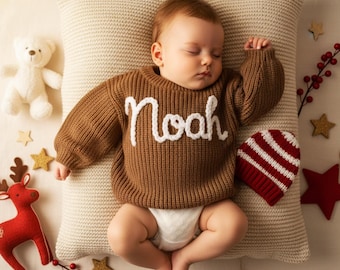 May include: A sleeping baby wearing a brown knit sweater with the name "Noah" in white script. The baby is lying on a beige pillow, surrounded by holiday decorations, including a red and white striped hat and a stuffed teddy bear.