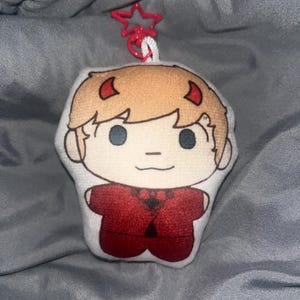 Puede incluir: Un llavero de peluche con un personaje de dibujos animados con cabello castaño claro, pequeños cuernos rojos y un traje rojo. El llavero tiene un clip rojo en forma de estrella y una cuerda blanca.