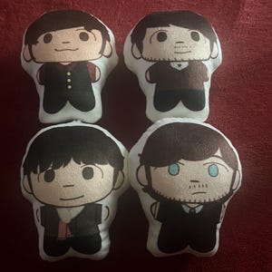 May include: Four plush dolls with cartoon-style faces and dark hair. Each doll has a different facial expression and is wearing a black suit. The dolls are arranged in a square on a maroon surface.