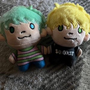 May include: Two plush dolls with embroidered features. The left doll has mint green hair, a pink and green striped shirt, and blue trousers. The right doll has yellow hair, green eyes, and a black shirt with the white text SideQuest printed on it.
