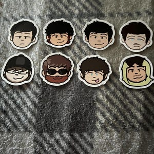 Puede incluir: Una colección de ocho pegatinas con ilustraciones de dibujos animados de caras masculinas con varios peinados y expresiones. Las pegatinas tienen un borde blanco y están dispuestas sobre una superficie gris texturizada.