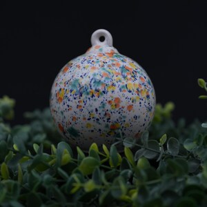 May include: A round, white ceramic ornament with a speckled design in blue, orange, and yellow. The ornament has a small loop at the top for hanging. The ornament is sitting on a bed of green foliage.