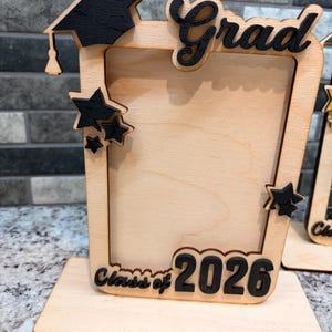 Op de afbeelding: Houten afstudeerfotolijst met een zwarte baret, sterren en het woord "Grad". De lijst bevat de tekst "Class of 2026" en is ontworpen om een foto in te plaatsen. De lijst is gemaakt van lichtgekleurd hout.