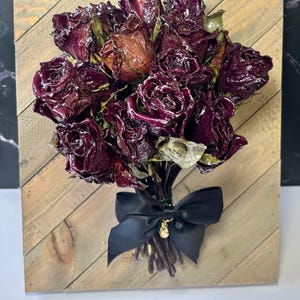 May include: A bouquet of dark red roses, preserved and arranged on a wooden plank. The roses are tied with a black satin bow, and a small gold charm hangs from the bow. The wooden plank has a natural wood grain pattern.