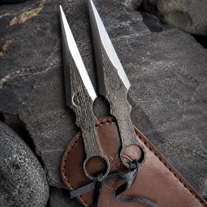 May include: Two metal throwing knives with sharp, silver blades and textured handles rest on a dark rock surface. The knives are paired with a brown leather sheath, secured with a black cord. The blades are approximately 15 cm long.