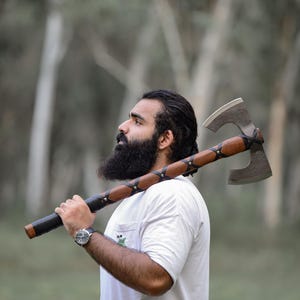 Könnte beinhalten: Eine handgefertigte Axt mit einem Holzgriff und einer Metallklinge. Der Griff ist mit Leder umwickelt und mit dekorativen Nähten versehen. Die Axt ist zum Hacken und Spalten von Holz bestimmt. Die Axt ist ein Werkzeug für Outdoor-Aktivitäten.