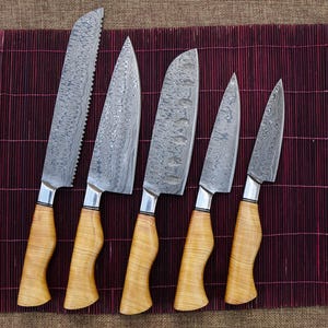 May include: A set of five Damascus steel knives with light wood handles. The blades have a unique patterned design. The handles are a light wood color and have a curved shape. The knives are arranged on a dark red bamboo mat.