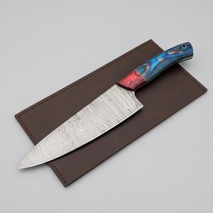 May include: A chef's knife with a patterned steel blade and a handle featuring red, blue, and black wood grain. The knife rests on a brown leather sheath, creating a striking contrast. This kitchen tool is designed for precision cutting.