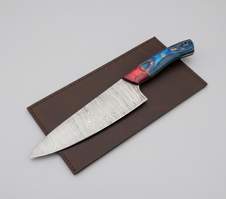 May include: A chef's knife with a patterned steel blade and a handle featuring red, blue, and black wood grain. The knife rests on a brown leather sheath, creating a striking contrast. This kitchen tool is designed for precision cutting.