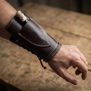 May include: A dark brown leather wrist cuff featuring a built-in sheath pocket containing a small knife with a light-colored handle. The cuff is secured with white stitching and a lace-up closure system that wraps around the forearm.