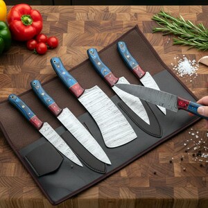 May include: A set of six kitchen knives with patterned blades and blue and red handles, displayed in a black leather knife roll. The knives are arranged on a wooden cutting board, with fresh vegetables in the background.