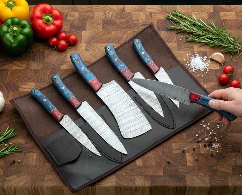 May include: A set of six kitchen knives with patterned blades and blue and red handles, displayed in a black leather knife roll. The knives are arranged on a wooden cutting board, with fresh vegetables in the background.