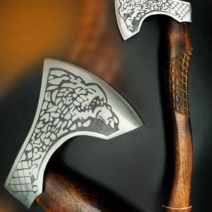 May include: Two axes with a detailed eagle design etched into the silver blades. The wooden handles are a dark brown with a carved diamond pattern near the blade. A leather strap is attached to the base of the handle.