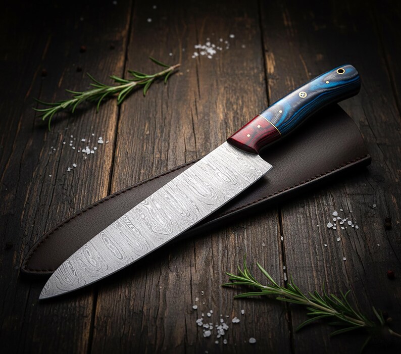 May include: A Damascus steel chef's knife with a patterned blade and a handle featuring blue, red, and gold accents. The knife rests in a brown leather sheath on a dark wooden surface, with rosemary sprigs and salt flakes scattered around.