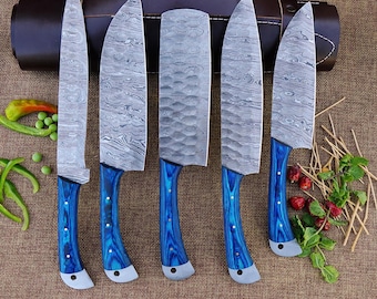 May include: A set of five Damascus steel kitchen knives with blue wooden handles. The blades have a unique swirling pattern. The knives are arranged in front of a brown leather knife roll, with some vegetables and herbs.