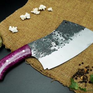 May include: A large, stainless steel cleaver with a purple and white handle. The blade has a hammered texture. The cleaver is on a burlap surface, with popcorn, rice, peppercorns, and a lemon slice.
