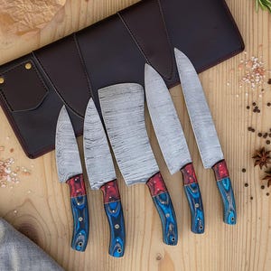 May include: A set of five kitchen knives with patterned steel blades and red and blue wooden handles. The knives are displayed on a wooden surface next to a brown leather knife case.