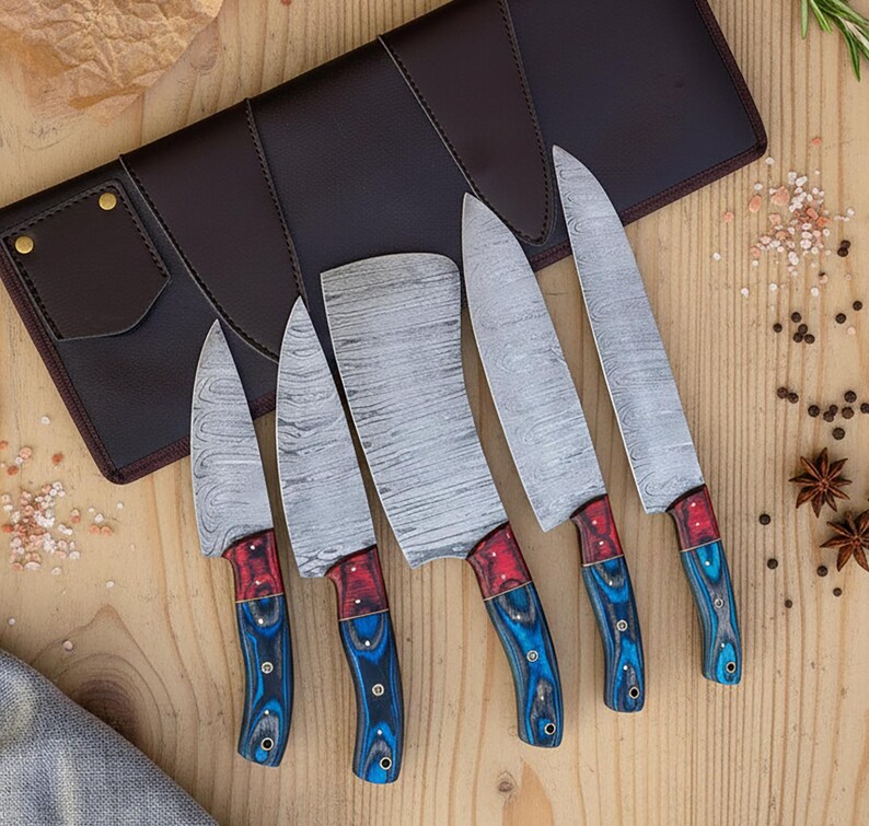 May include: A set of five kitchen knives with patterned steel blades and red and blue wooden handles. The knives are displayed on a wooden surface next to a brown leather knife case.