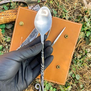 May include: A hand-forged metal spoon with a twisted handle and a decorative loop at the end. The spoon is held against a backdrop of a leather sheath and other metal tools, suggesting a rustic or artisanal theme.