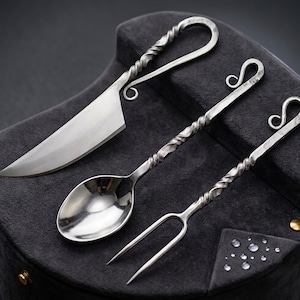 May include: A set of three hand-forged metal utensils including a knife, a spoon, and a two-pronged fork. Each piece features a twisted handle design with a curled end. The metal has a dark, textured finish with polished, reflective surfaces.