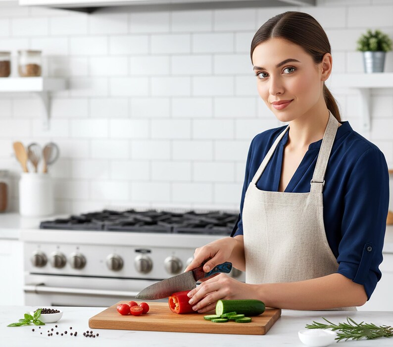 May include: A woman in a blue shirt and beige apron is cutting vegetables on a wooden cutting board in a kitchen. Red bell pepper, tomatoes, and cucumber slices are visible. A stainless steel knife is in use.