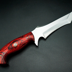 May include: A custom knife with a polished steel blade and a vibrant red and black wooden handle. The handle features a decorative brass inlay. The knife is designed with a curved blade and a pointed tip.