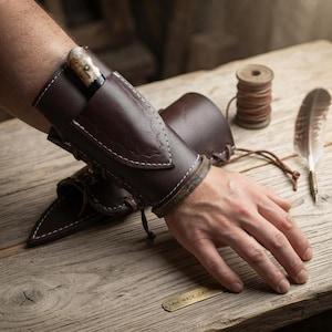 May include: A dark brown leather bracer featuring white contrast stitching, a lace-up closure, and an integrated sheath holding a small knife with a light-colored handle. The leather has a smooth finish with decorative embossed edge detailing.
