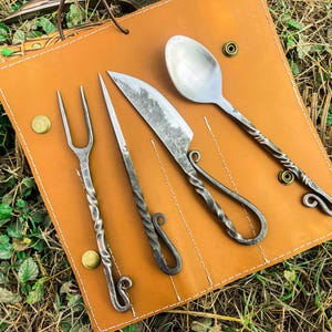 May include: A handcrafted, rustic cutlery set includes a fork, knife, spoon, and a small knife, all with twisted handles and decorative swirls. The set is displayed on a brown leather carrying case.