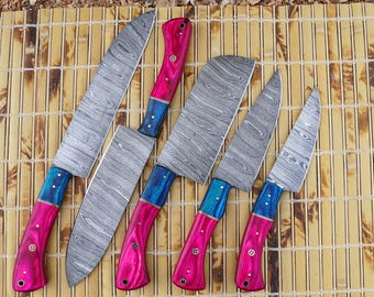 Pode incluir: Um conjunto de seis facas de cozinha com lâminas de aço estampadas e cabos vibrantes. Os cabos são uma combinação de rosa e azul, com rebites decorativos. As facas estão dispostas sobre uma esteira de bambu.