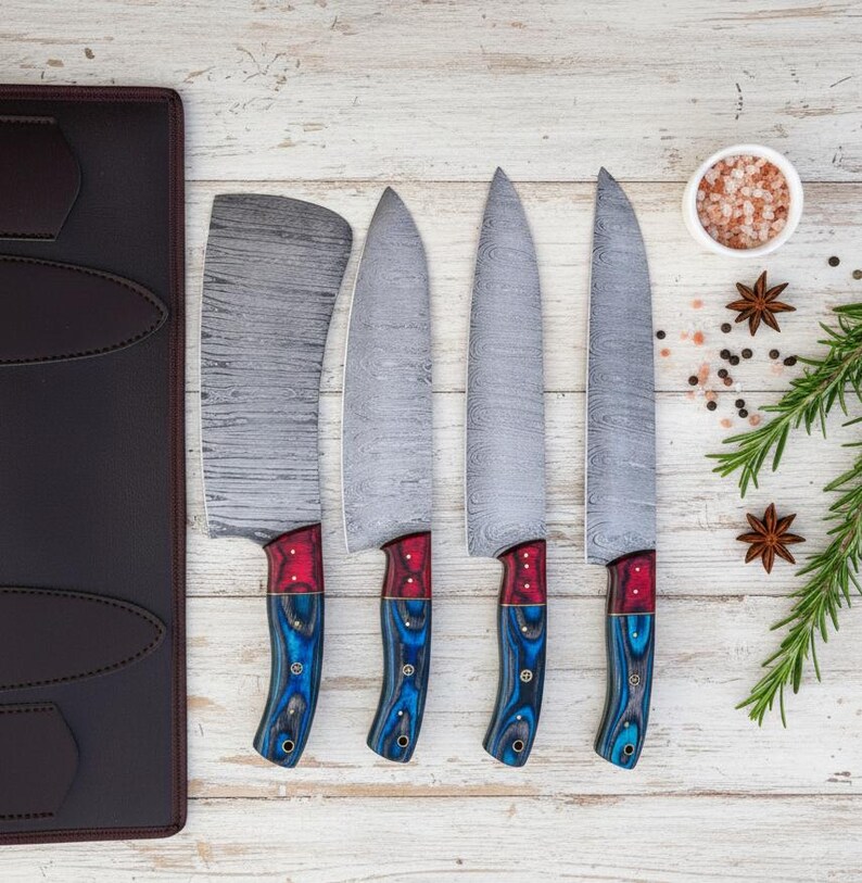 May include: A set of four kitchen knives with patterned steel blades and red and blue handles. The knives are arranged on a light wood surface next to a dark brown leather knife case. A small bowl of pink salt and spices are also present.